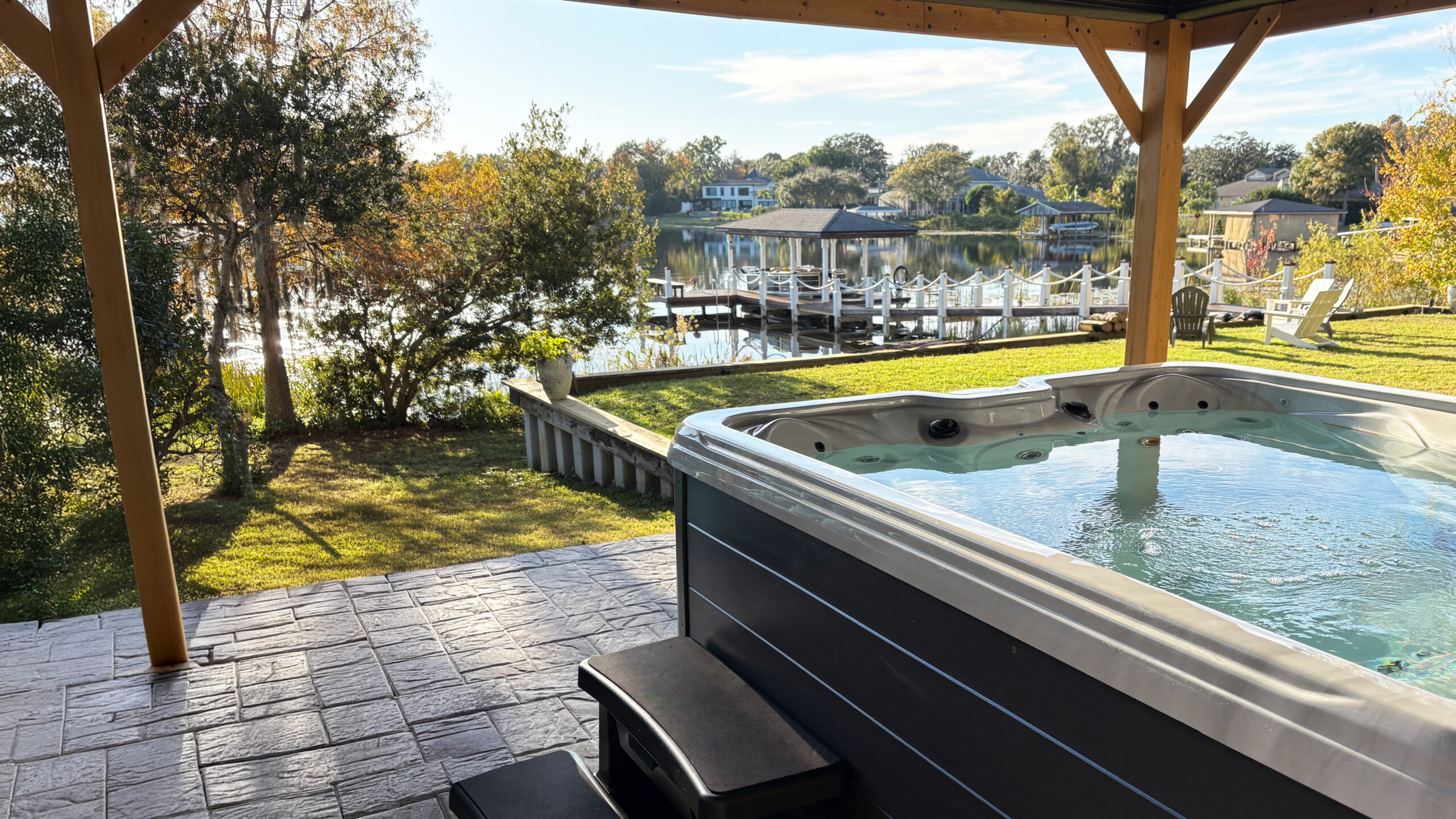 One of Whisper Outdoor's 110V hot tubs on a covered patio overlooking a lakefront backyard with a dock and water view.